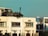 A modern, multi-story apartment building with a rooftop garden filled with greenery. The facade is slightly weathered with visible air conditioning units. Large windows allow natural light into the rooms. The sky in the background is clear and blue.