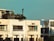 A modern, multi-story apartment building with a rooftop garden filled with greenery. The facade is slightly weathered with visible air conditioning units. Large windows allow natural light into the rooms. The sky in the background is clear and blue.