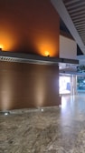 A modern interior space featuring a polished stone floor and wooden walls illuminated by warm, recessed lighting. A grid-like ceiling structure adds architectural interest, and an exterior view of trees is partially visible through glass doors or windows.