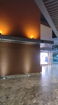 A modern interior space featuring a polished stone floor and wooden walls illuminated by warm, recessed lighting. A grid-like ceiling structure adds architectural interest, and an exterior view of trees is partially visible through glass doors or windows.
