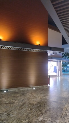 A modern interior space featuring a polished stone floor and wooden walls illuminated by warm, recessed lighting. A grid-like ceiling structure adds architectural interest, and an exterior view of trees is partially visible through glass doors or windows.