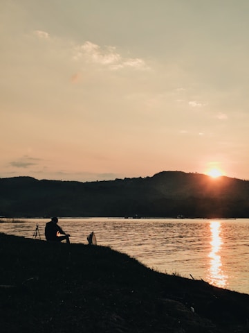 A serene landscape featuring a silhouette of a person sitting by a riverbank with a calm body of water and mountains in the background. The sun is setting, casting a warm glow across the sky and reflecting on the river. A tripod is placed nearby, suggesting photography or observation.