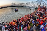 A large crowd of people gathers by the riverside during a vibrant cultural or religious festival. Many individuals are dressed in bright, colorful clothing, with some holding offerings or baskets. In the background, a prominent steel bridge spans the river, and boats can be seen on the water. The scene captures a sense of communal celebration and tradition.