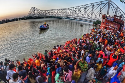 A large crowd of people gathers by the riverside during a vibrant cultural or religious festival. Many individuals are dressed in bright, colorful clothing, with some holding offerings or baskets. In the background, a prominent steel bridge spans the river, and boats can be seen on the water. The scene captures a sense of communal celebration and tradition.