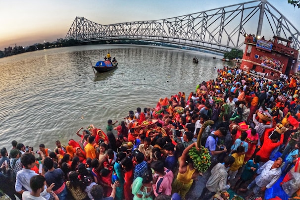 A large crowd of people gathers by the riverside during a vibrant cultural or religious festival. Many individuals are dressed in bright, colorful clothing, with some holding offerings or baskets. In the background, a prominent steel bridge spans the river, and boats can be seen on the water. The scene captures a sense of communal celebration and tradition.