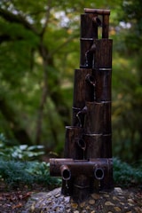 A bamboo water feature consisting of several dark bamboo poles stacked vertically and horizontally, with water flowing gently from the top down each level. The background is filled with lush green foliage, creating a serene and natural setting.