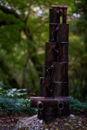A bamboo water feature consisting of several dark bamboo poles stacked vertically and horizontally, with water flowing gently from the top down each level. The background is filled with lush green foliage, creating a serene and natural setting.