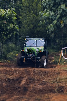 A rugged off-road vehicle is navigating through a muddy terrain surrounded by dense greenery. The vehicle has large tires and is outfitted with off-road modifications, including a winch. Its bright green color stands out against the earthy brown of the mud and the lush green foliage.
