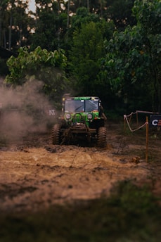 A rugged, off-road vehicle is navigating a muddy terrain surrounded by dense greenery. Dust is kicked up by the tires, creating a dynamic scene of adventure and exploration. The vehicle is prominently marked with decals and appears to be in motion, showcasing its off-road capabilities.