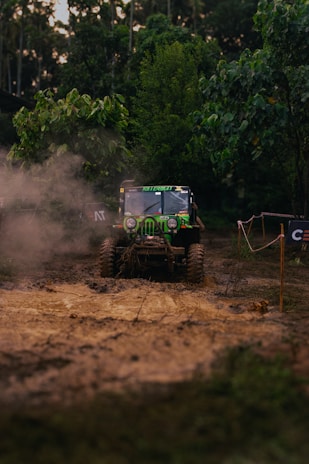 A rugged, off-road vehicle is navigating a muddy terrain surrounded by dense greenery. Dust is kicked up by the tires, creating a dynamic scene of adventure and exploration. The vehicle is prominently marked with decals and appears to be in motion, showcasing its off-road capabilities.