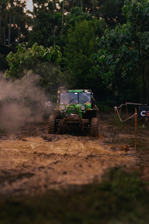 A rugged, off-road vehicle is navigating a muddy terrain surrounded by dense greenery. Dust is kicked up by the tires, creating a dynamic scene of adventure and exploration. The vehicle is prominently marked with decals and appears to be in motion, showcasing its off-road capabilities.