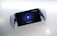 A handheld gaming device is positioned against a white background. The device features a screen displaying a PlayStation logo with the prompt 'To unlock, press the PS button.' It is framed by white controllers on both sides, resembling a hybrid of a tablet and a console.