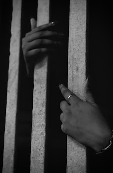 A pair of hands grip metal bars, suggesting confinement or imprisonment. The lighting is dim, emphasizing the hands and creating shadows.