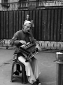 A man is seated on a plastic stool playing a harmonium in an outdoor setting. He wears a cap, glasses, and casual clothing. In the background, a person is standing near a wrought iron fence and wooden wall. The image is in black and white, adding a classic or nostalgic mood.