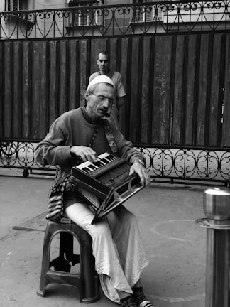 A man is seated on a plastic stool playing a harmonium in an outdoor setting. He wears a cap, glasses, and casual clothing. In the background, a person is standing near a wrought iron fence and wooden wall. The image is in black and white, adding a classic or nostalgic mood.