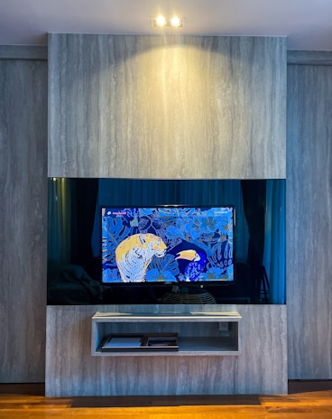 A modern interior with a mounted TV displaying a vibrant, colorful artwork. The background is blue with floral patterns and features a yellow tiger and a toucan. The TV is framed by a sleek, light gray panel with a shelf underneath holding a few items. Warm lighting illuminates the setup, creating a cozy atmosphere.
