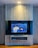 A modern interior with a mounted TV displaying a vibrant, colorful artwork. The background is blue with floral patterns and features a yellow tiger and a toucan. The TV is framed by a sleek, light gray panel with a shelf underneath holding a few items. Warm lighting illuminates the setup, creating a cozy atmosphere.