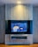 A modern interior with a mounted TV displaying a vibrant, colorful artwork. The background is blue with floral patterns and features a yellow tiger and a toucan. The TV is framed by a sleek, light gray panel with a shelf underneath holding a few items. Warm lighting illuminates the setup, creating a cozy atmosphere.