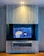 A modern interior with a mounted TV displaying a vibrant, colorful artwork. The background is blue with floral patterns and features a yellow tiger and a toucan. The TV is framed by a sleek, light gray panel with a shelf underneath holding a few items. Warm lighting illuminates the setup, creating a cozy atmosphere.