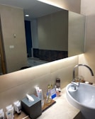 A well-organized bathroom counter with a white ceramic sink and a silver faucet. Toiletries like toothpaste, toothbrushes, and other personal care items are neatly arranged on the counter. The countertop is made of light-colored stone, and a large mirror occupies the wall above the sink. The warm lighting creates a cozy atmosphere, reflecting off beige tiled walls.