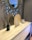 A modern interior design with a large decorative mirror featuring a sunburst pattern. A light wooden console table displays a black reed diffuser, a circular decorative wooden plate, and a clear box with seashells. The setting conveys a clean and contemporary aesthetic.