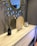 A modern interior design with a large decorative mirror featuring a sunburst pattern. A light wooden console table displays a black reed diffuser, a circular decorative wooden plate, and a clear box with seashells. The setting conveys a clean and contemporary aesthetic.