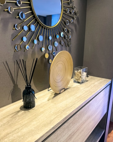 A modern interior design with a large decorative mirror featuring a sunburst pattern. A light wooden console table displays a black reed diffuser, a circular decorative wooden plate, and a clear box with seashells. The setting conveys a clean and contemporary aesthetic.