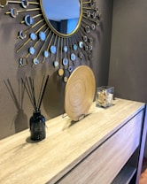 A modern interior design with a large decorative mirror featuring a sunburst pattern. A light wooden console table displays a black reed diffuser, a circular decorative wooden plate, and a clear box with seashells. The setting conveys a clean and contemporary aesthetic.