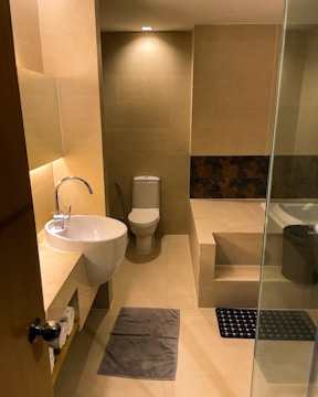 A modern bathroom with beige tiled walls and floor. It features a wall-mounted round sink with a curved faucet, a toilet positioned at the far end, and a raised platform holding a bathtub partially covered by frosted glass. The lighting is warm, enhancing the neutral tones. A small mirror hangs above the sink, and there are rolled towels on a shelf below.