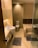 A modern bathroom with beige tiled walls and floor. It features a wall-mounted round sink with a curved faucet, a toilet positioned at the far end, and a raised platform holding a bathtub partially covered by frosted glass. The lighting is warm, enhancing the neutral tones. A small mirror hangs above the sink, and there are rolled towels on a shelf below.