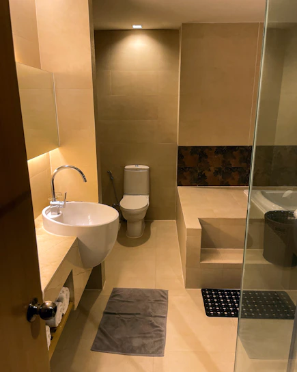 A modern bathroom with beige tiled walls and floor. It features a wall-mounted round sink with a curved faucet, a toilet positioned at the far end, and a raised platform holding a bathtub partially covered by frosted glass. The lighting is warm, enhancing the neutral tones. A small mirror hangs above the sink, and there are rolled towels on a shelf below.