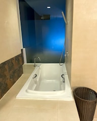 A modern bathroom featuring a white built-in bathtub with chrome fixtures including a handheld shower head attached to the wall. The bathtub is set against a glass wall with a blue hue, providing a contemporary look. Adjacent to the bathtub is a dark wicker basket for storage. The walls and floor are covered with beige tiles, adding a warm tone to the space.