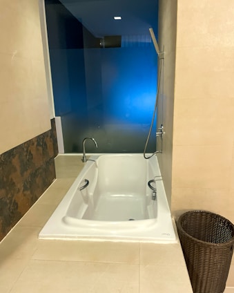 A modern bathroom featuring a white built-in bathtub with chrome fixtures including a handheld shower head attached to the wall. The bathtub is set against a glass wall with a blue hue, providing a contemporary look. Adjacent to the bathtub is a dark wicker basket for storage. The walls and floor are covered with beige tiles, adding a warm tone to the space.