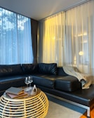 A modern living room with a black leather sectional sofa adorned with a beige throw blanket. The room features large windows with sheer curtains, allowing natural light to filter in, creating a warm atmosphere. In front of the sofa, there is a round wicker coffee table with a glass top, on which two empty wine glasses and some books rest.