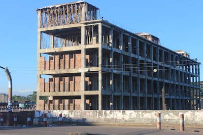 A partially constructed multi-story building is visible, with exposed brick walls and concrete beams. The upper floors have wooden scaffolding and reinforcing rods protruding. The scene is set on a clear day with a blue sky, and the building is surrounded by a road with barriers.