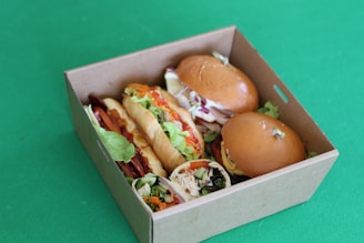 A cardboard box filled with various types of sandwiches and wraps. The box contains two round bread sandwiches with lettuce, a sub-style sandwich, and a wrap with visible ingredients like lettuce and other veggies. The background is a solid green surface.