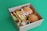 A cardboard box filled with various types of sandwiches and wraps. The box contains two round bread sandwiches with lettuce, a sub-style sandwich, and a wrap with visible ingredients like lettuce and other veggies. The background is a solid green surface.
