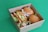 A cardboard box filled with various types of sandwiches and wraps. The box contains two round bread sandwiches with lettuce, a sub-style sandwich, and a wrap with visible ingredients like lettuce and other veggies. The background is a solid green surface.