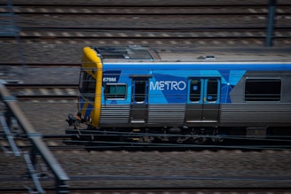 A metro train is moving quickly along a set of railway tracks. The train is predominantly silver with a blue and yellow design, featuring the word 'METRO' on its side. The tracks are surrounded by blurred motion, indicating speed.