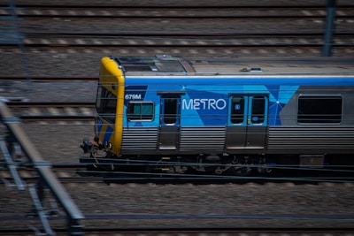A metro train is moving quickly along a set of railway tracks. The train is predominantly silver with a blue and yellow design, featuring the word 'METRO' on its side. The tracks are surrounded by blurred motion, indicating speed.