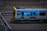 A metro train is moving quickly along a set of railway tracks. The train is predominantly silver with a blue and yellow design, featuring the word 'METRO' on its side. The tracks are surrounded by blurred motion, indicating speed.