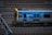 A metro train is moving quickly along a set of railway tracks. The train is predominantly silver with a blue and yellow design, featuring the word 'METRO' on its side. The tracks are surrounded by blurred motion, indicating speed.