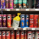 A variety of snack containers are neatly arranged on supermarket shelves. The products include cans of Pringles chips in different flavors such as BBQ, Original, Salt & Vinegar, Habanero, and Scorchin' Sour Cream & Onion. A yellow bag of chips is placed between the cans. Also visible are Doritos Minis Nacho Cheese containers. Prices are displayed on tags below each row.