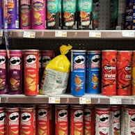 A variety of snack containers are neatly arranged on supermarket shelves. The products include cans of Pringles chips in different flavors such as BBQ, Original, Salt & Vinegar, Habanero, and Scorchin' Sour Cream & Onion. A yellow bag of chips is placed between the cans. Also visible are Doritos Minis Nacho Cheese containers. Prices are displayed on tags below each row.