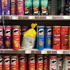 A variety of snack containers are neatly arranged on supermarket shelves. The products include cans of Pringles chips in different flavors such as BBQ, Original, Salt & Vinegar, Habanero, and Scorchin' Sour Cream & Onion. A yellow bag of chips is placed between the cans. Also visible are Doritos Minis Nacho Cheese containers. Prices are displayed on tags below each row.