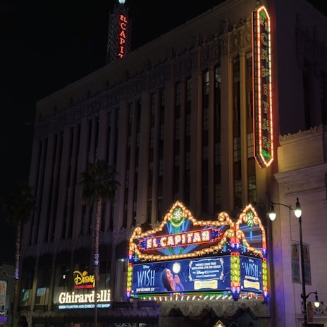 A theater marquee brightly illuminated at night, featuring neon lights and a vertical sign with the name 'El Capitan'. The marquee advertises a Disney movie 'Wish' with colorful lights adding vibrancy to the scene. Surrounding buildings include one with a Disney Studio Store and a Ghirardelli Soda Fountain and Chocolate Shop. Palm trees are visible alongside tall, classical architecture.