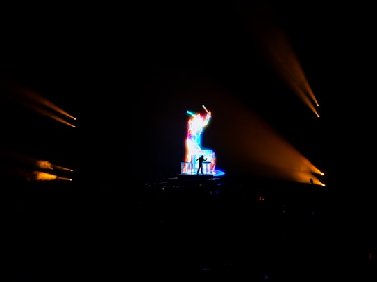 A vibrant, multicolored light display highlights a silhouette of a person standing on a stage. The person appears to be playing a musical instrument, possibly a guitar. The bright colors stand out dramatically against the dark background, with beams of light visible.
