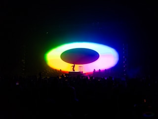A colorful, illuminated stage setup with vibrant lights forming a large oval shape. A silhouette of a performer and a piece of equipment are visible in the center, surrounded by an audience in darkness.