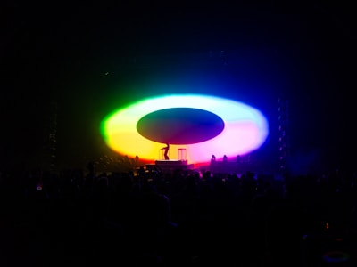 A colorful, illuminated stage setup with vibrant lights forming a large oval shape. A silhouette of a performer and a piece of equipment are visible in the center, surrounded by an audience in darkness.