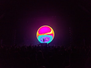 A vibrant, colorful circular screen dominates the center of the image, displaying swirling psychedelic patterns in hues of pink, yellow, and blue. In the foreground, the silhouette of a crowd is visible, suggesting the setting of a concert or live event.